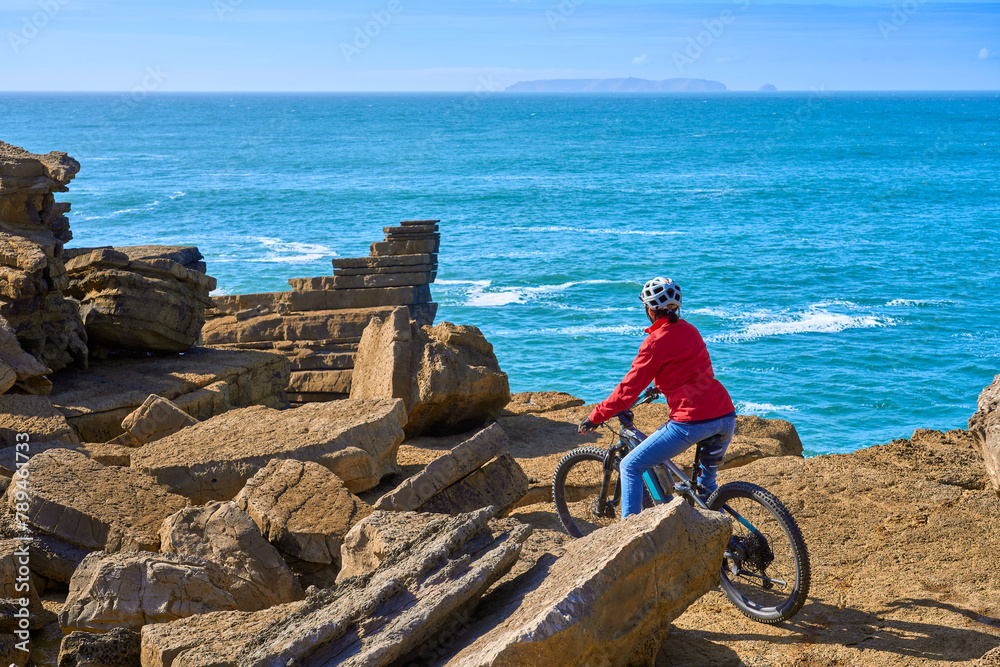 Wall mural Brave senior woman riding her electric mountain bike on the rocky cliffs of Peniche at the western atlantic coast of Portugal