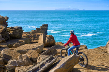 Brave senior woman riding her electric mountain bike on the rocky cliffs of Peniche at the western atlantic coast of Portugal - 789461733