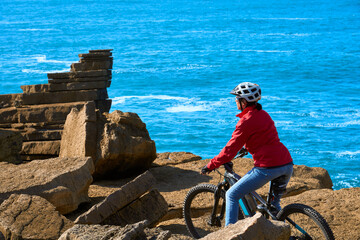 Brave senior woman riding her electric mountain bike on the rocky cliffs of Peniche at the western atlantic coast of Portugal - 789461781