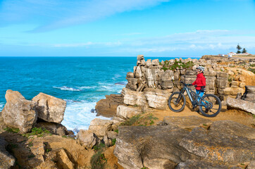 Brave senior woman riding her electric mountain bike on the rocky cliffs of Peniche at the western atlantic coast of Portugal - 789462150
