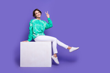Full size photo of attractive young woman sit white cube show v-sign dressed stylish green clothes isolated on violet color background