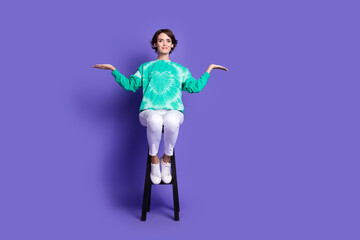 Full size photo of attractive woman sit chair compare hold empty space dressed stylish green clothes isolated on violet color background
