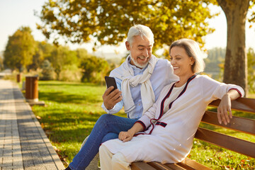 Smiling happy senior active couple man and woman sitting on the bench in a public park using mobile...