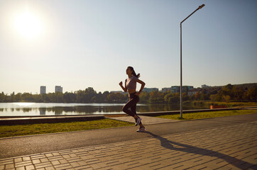 Confident motivated female athlete having outdoor jogging workout. Woman runner in good physical shape running along waterfront, with beautiful city park view in background. Sport, fitness concept - obrazy, fototapety, plakaty