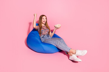 Full length photo of lovely teen lady sit bean bag eat popcorn movie winner dressed stylish print clothes isolated on pink color background