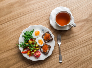 Delicious breakfast, brunch, snack - boiled egg, fresh vegetable salad, red caviar toasts and tea on a wooden background, top view
