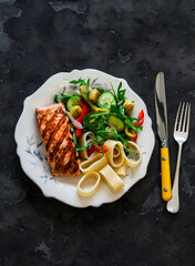 Balanced lunch - grilled salmon, pasta and fresh vegetable salad on a dark background, top view
