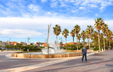 Obraz premium Man walking in city. People on a walk along path near Fountain. Fountain in form of sailboat. Fuente del Barco de Agua de la Playa Malvarrosa in Valencia, Spain. Male tourist walking along sidewalk