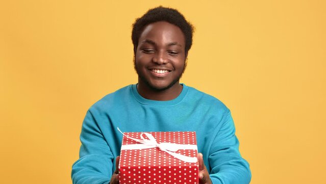 Cute African American young man giving gift box to camera, portrait in studio with orange background