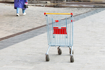 an empty shopping cart stands on the sidewalk. the concept of people lacking money for purchases. economic crisis. Shopping cart. leaving unrecognizable person - obrazy, fototapety, plakaty