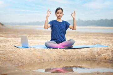 indian teenage girl sitting in a easy pose doing gyana mudra hatha yoga by learning on laptop at a...