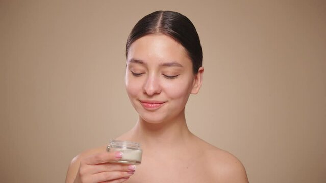 Funny happy attractive young woman applies skin care cream to her face, looks at the camera, applies moisturizing facelift cream. Slow motion