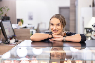 A cute female cafe owner is leaning against a glass display case with cakes and smiling.. Young female barista standing behind the bar in cafe smiling - 789501732