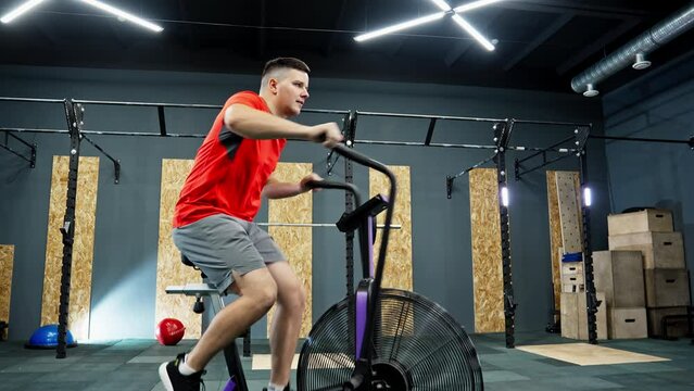 Young man riding stationary bike indoors. Sportsman spinning on cardio machine. Functional training. Gym bike. Dolly zoom in. Gym is well lit and has modern design. 4K, UHD