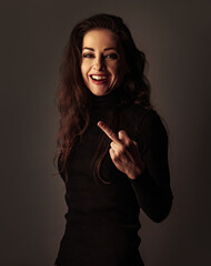 Fuck you, whiner. Beautiful happy laughing woman showing fuck off sign by middle finger in black clothing on dark studio background on empty space. Closeup