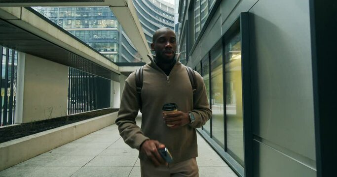 A man uses a mobile phone and drinks coffee from a disposable cup walking along a pedestrian city street