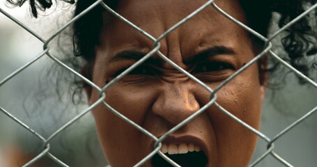 One upset young black woman yelling behind metal fence barrier looking at camera grimacing in anger...