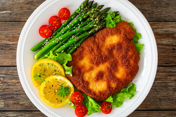 Crispy breaded fried pork chop, green asparagus and fresh vegetables on wooden table
