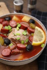 Meat solyanka soup with thin dry smoked sausages in bowl on wooden board, closeup