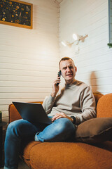 Attractive man sitting on the sofa at home and talking on the phone, working on a laptop online, freelancer modern lifestyle. - obrazy, fototapety, plakaty