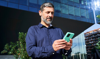 Cheerful mature businessman using phone to send text messages to be connected, standing outside of a corporate district. Copy space.