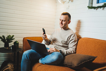 Attractive man sitting on the sofa at home and talking on the phone, working on a laptop online, freelancer modern lifestyle. - obrazy, fototapety, plakaty