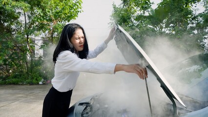 Worried woman in white blouse stands by car with smoking engine, squinting from smoke, in verdant area. Concept of taking care of car insurance after an accident. - obrazy, fototapety, plakaty