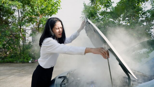 Worried woman in white blouse stands by car with smoking engine, squinting from smoke, in verdant area. Concept of taking care of car insurance after an accident.