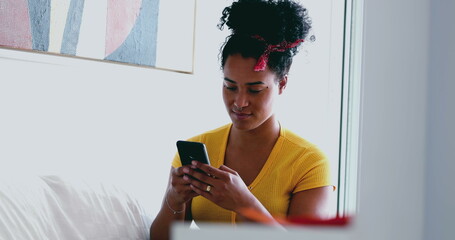 Young African American woman texting message at home seated at home sofa. One candid black latina...