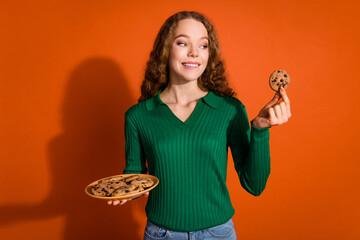 Photo of hungry unsure lady dressed green shirt biting lip enjoying cookies empty space isolated orange color background