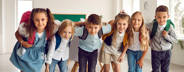 Portrait of cheerful group of elementary school students with backpacks on their shoulders in classroom. Cute little boys and girls standing in row, hugging and smiling at camera. Web banner.