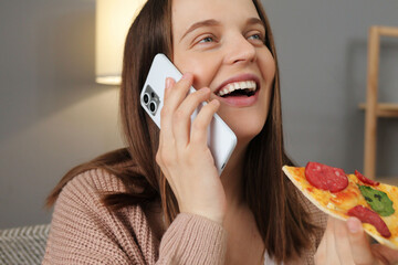 Caucasian woman has phone conversation while eating tasty pizza holding smartphone laughing happily...