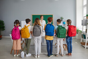 Small group of children in casual clothes gets acquainted with smiling teacher of individual classes. Teenagers with backpacks on their backs lined up in front of their teacher in the classroom.