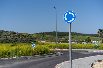 A solitary roundabout traffic sign against a backdrop of a vibrant yellow field and clear skies. Traffic sign with a circular arrow pointing the way for roundabout navigation.
