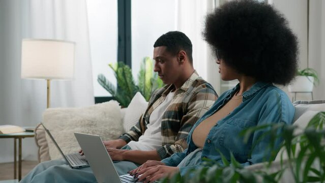 Gadget video game addict family computers addicted African American couple on couch at home ignoring using two laptops sad African American woman wife girlfriend looking on husband man exhale stress