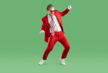 Fototapeta na wymiar New Year's fun. Cool stylish man in fashionable red suit is having fun dancing at New Year's party. Full length of cheerful man in suit with tie and sunglasses hanging out on green background.