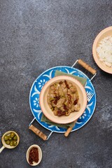 A hot dish, stewed beef with dried tomatoes and olives in a clay bowl on a blue meallic tray on a dark concrete background. Beef recipes.