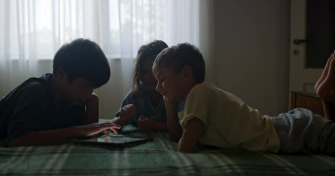Three children engaged in playful interaction on a bed by a digital tablet at dusk