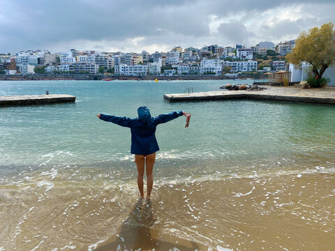 A girl in rainy jacket raises her arms in the charming Agios Nikolaos.