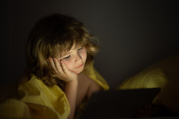 Kid playing game on tablet in bed at night. Kids with social media. Child lying in bed playing a tablet in dark room, light under blacket. Close up of kid watching cartoons on tablet.