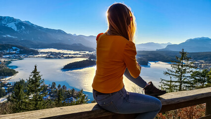 Fototapeta na wymiar Woman sitting on railing of Taborhoehe viewing platform in Carinthia, Austria, Europe. Surrounded by high snow capped Austrian Alps mountains. lake surface is frozen. Alpine Landscape in frosty winter