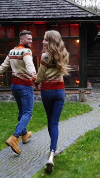 Young couple in the village. Handsome man and beautiful girl in Christmas sweaters are running in the yard of cottage. Romantic couple enjoying life outdoors. Vertical video