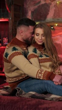 Lovely couple in romantic bedroom. Beautiful young woman and her boyfriend in Christmas sweaters sitting on bed in cosy decorated room. Vertical video