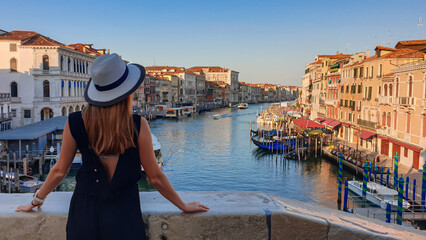 Tourist woman standing on top of famous Rialto bridge overlooking the Canal Grande in Venice, Veneto, Northern Italy, Europe. Female model is wearing black dress. Romantic luxury summer vacation