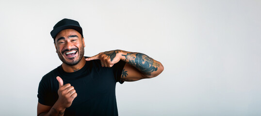 A latino man with a black shirt and a cap is smiling and pointing to his mouth. He has a tattoo on his arm, isolated on white 