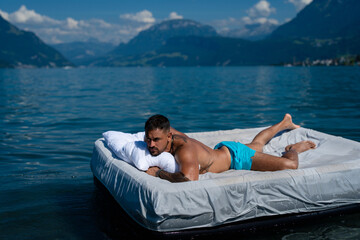 Tourist rest at the summer lake. Summer trip in nature. Peaceful water. Muscular sexy man lying on cozy comfort Mattresses. Sleep and dreams concept. Sexy man floating on the Bed with blanket.