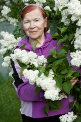 Outdoor close-up portrait of old senior woman. Beautiful elderly woman smiling against background of blooming white lilacs in spring park.