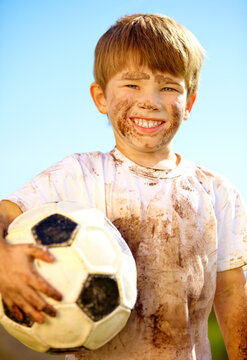 Portrait, smile and kid with soccer ball outdoor for sports, exercise or game in summer. Face, boy and happy child with football with mud, messy or dirty for recreation in Switzerland on blue sky