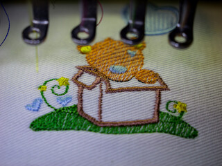 Machine embroidery on white fabric. Little bear sitting in a box on a green meadow. Selective focus.