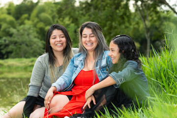 three latina girls, friends together laughing loudly sitting on the lawn next to the fishing lake